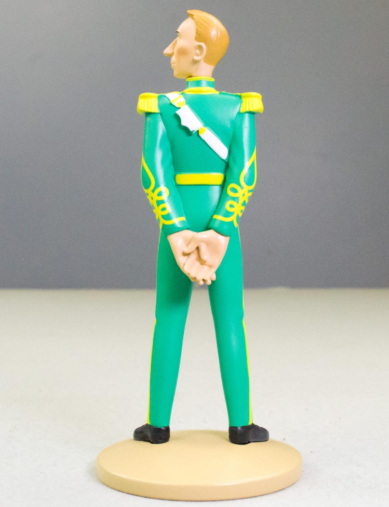 May include: A figurine of a man in a green uniform with yellow accents and a white sash. The figure has blond hair and is standing on a tan circular base. The uniform features decorative yellow scrollwork.