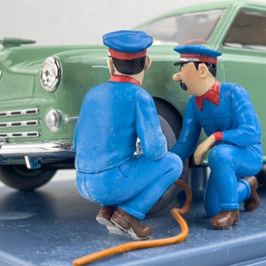 Pu&ograve; includere: Una scena in miniatura con due figure in uniforme blu e berretto, intente a lavorare su un'auto verde classica. Le figure sono vicino allo pneumatico, collegate da un tubo arancione. L'auto ha dettagli cromati.