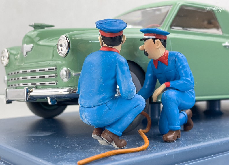Pu&ograve; includere: Una scena in miniatura con due figure in uniforme blu e berretto, intente a lavorare su un'auto verde classica. Le figure sono vicino allo pneumatico, collegate da un tubo arancione. L'auto ha dettagli cromati.