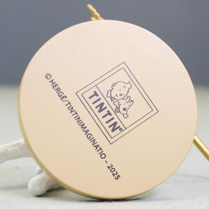 Puede incluir: Primer plano de la base de una figura circular beige. La figura presenta el logotipo "Tint&iacute;n" y el texto "&copy; HERGE/TINTINIMAGINATIO - 2025". Tambi&eacute;n se ven un objeto blanco y una varilla dorada.