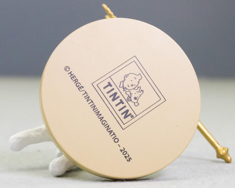Puede incluir: Primer plano de la base de una figura circular beige. La figura presenta el logotipo "Tint&iacute;n" y el texto "&copy; HERGE/TINTINIMAGINATIO - 2025". Tambi&eacute;n se ven un objeto blanco y una varilla dorada.