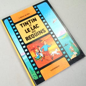 May include: A vintage hardcover book titled "Tintin and the Lake of Sharks" in French. The cover features a film strip design with illustrations of Tintin, Snowy, and other characters running. The book has a yellow and blue color scheme.