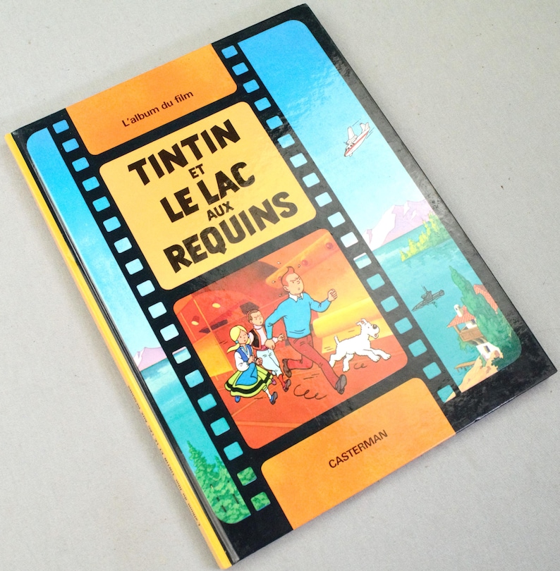 May include: A vintage hardcover book titled "Tintin and the Lake of Sharks" in French. The cover features a film strip design with illustrations of Tintin, Snowy, and other characters running. The book has a yellow and blue color scheme.