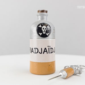 Könnte beinhalten: Eine silberne und gelbe Flasche mit Totenkopf und Knochen und dem Wort "RADJAIDJ" auf dem Etikett. Neben der Flasche liegt eine silberne Spritze mit gelber Spitze.