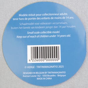 May include: A circular blue label with white text and a barcode. The text includes warnings about keeping the item out of reach of children under 14 years old. The label also states it is a small scale collectible model.