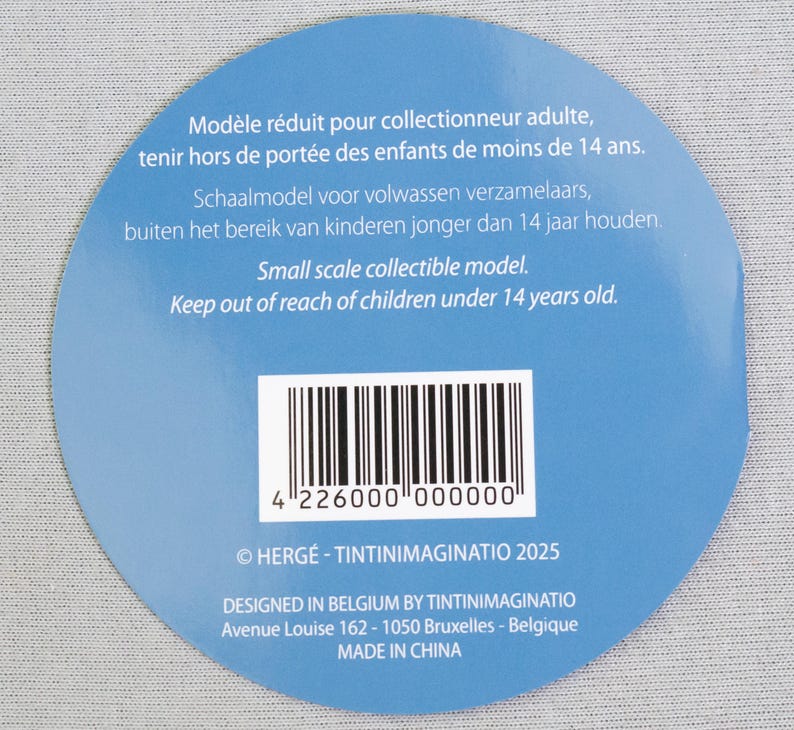 May include: A circular blue label with white text and a barcode. The text includes warnings about keeping the item out of reach of children under 14 years old. The label also states it is a small scale collectible model.