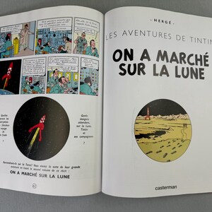Puede incluir: Libro abierto con ilustraciones de c&oacute;mic de un lanzamiento de cohete y astronautas. El t&iacute;tulo "ON A MARCH&Eacute; SUR LA LUNE" es visible. Es una aventura de Tint&iacute;n, con el logo de la editorial.