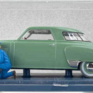 Pu&ograve; includere: Un modello di auto d'epoca verde con pneumatici a fascia bianca e dettagli cromati, esposto in una teca trasparente. Due figure in miniatura in uniforme blu sembrano lavorare sull'auto. Il modello &egrave; montato su una base blu scuro.