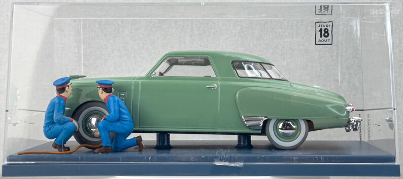Pu&ograve; includere: Un modello di auto d'epoca verde con pneumatici a fascia bianca e dettagli cromati, esposto in una teca trasparente. Due figure in miniatura in uniforme blu sembrano lavorare sull'auto. Il modello &egrave; montato su una base blu scuro.