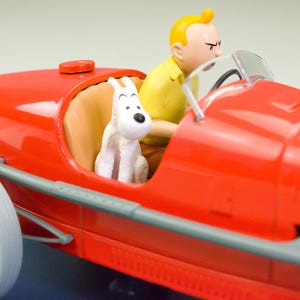 Pu&ograve; includere: Un'auto da corsa giocattolo rossa con un pilota e una statuina di cane bianco. Il pilota indossa una camicia gialla e il cane &egrave; seduto sul sedile del passeggero. L'auto ha pneumatici bianchi e raggi neri.