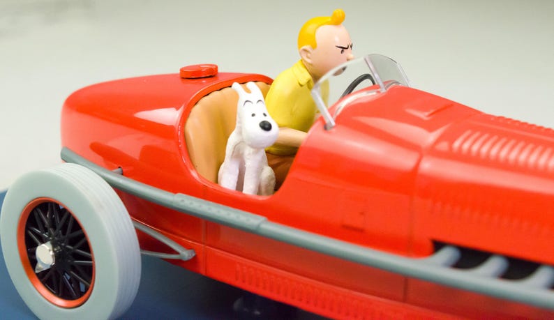 Pu&ograve; includere: Un'auto da corsa giocattolo rossa con un pilota e una statuina di cane bianco. Il pilota indossa una camicia gialla e il cane &egrave; seduto sul sedile del passeggero. L'auto ha pneumatici bianchi e raggi neri.