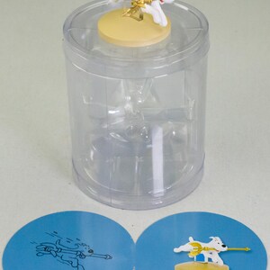 Puede incluir: Una figura de perro blanco con una lanza dorada y alas, sobre una base amarilla, dentro de un recipiente de pl&aacute;stico transparente. Dos tarjetas circulares azules con ilustraciones de perros est&aacute;n en la parte inferior.