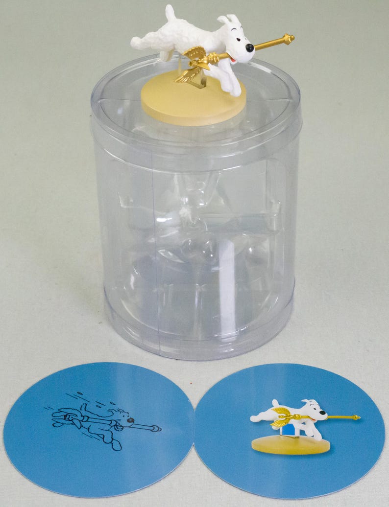 Puede incluir: Una figura de perro blanco con una lanza dorada y alas, sobre una base amarilla, dentro de un recipiente de pl&aacute;stico transparente. Dos tarjetas circulares azules con ilustraciones de perros est&aacute;n en la parte inferior.