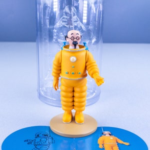 May include: A yellow astronaut figurine with a blue helmet and a white face. The figurine is standing on a round base with a blue background. The figurine is in a clear plastic container.