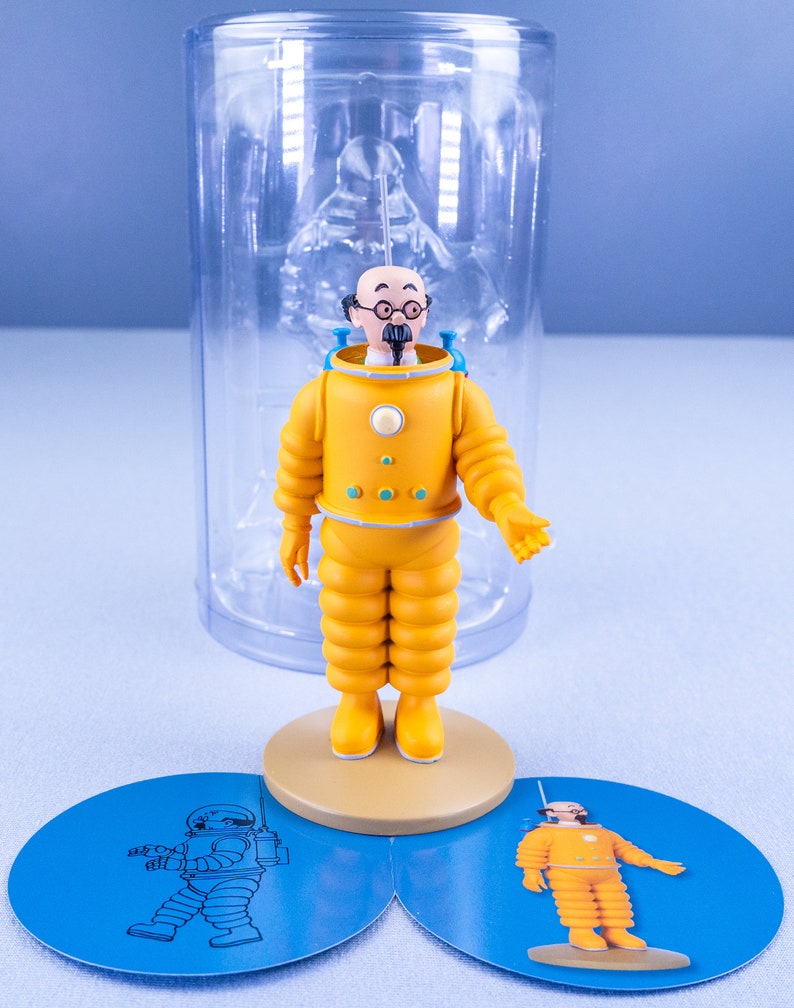 May include: A yellow astronaut figurine with a blue helmet and a white face. The figurine is standing on a round base with a blue background. The figurine is in a clear plastic container.