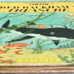 May include: A vintage hardcover book titled "The Adventures of Tintin: Red Rackham's Treasure." The cover features an illustration of a black and white shark-shaped submarine, red fish, and underwater plants against a teal background. The book has a red spine.