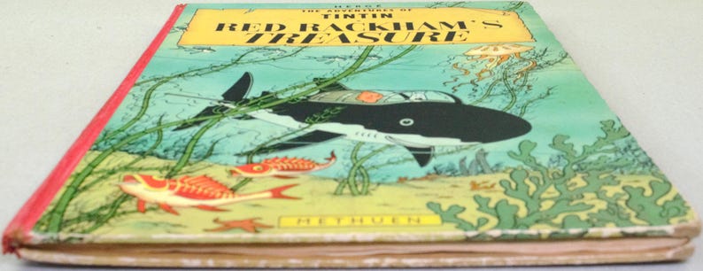 May include: A vintage hardcover book titled "The Adventures of Tintin: Red Rackham's Treasure." The cover features an illustration of a black and white shark-shaped submarine, red fish, and underwater plants against a teal background. The book has a red spine.