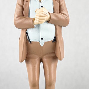 May include: A painted figurine of a man with a mustache wearing a green hat, a light blue shirt, and a tan suit. The figure stands on a circular base with hands clasped together. The text TINTINOLOGY is visible in the upper right corner.