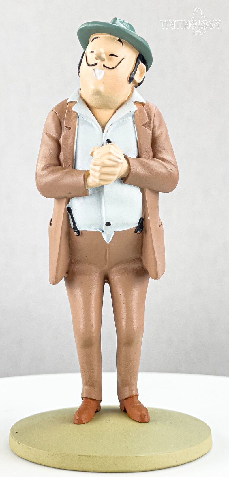 May include: A painted figurine of a man with a mustache wearing a green hat, a light blue shirt, and a tan suit. The figure stands on a circular base with hands clasped together. The text TINTINOLOGY is visible in the upper right corner.