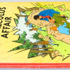 May include: A vintage copy of "The Calculus Affair" by Herg&eacute;, featuring a yellow cover with the title in bold black letters. The cover art depicts a dynamic scene with characters in action, set against a backdrop of trees and explosions. The book has a red spine.