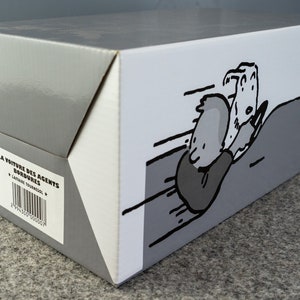 Puede incluir: Una caja de cart&oacute;n gris y blanca con una ilustraci&oacute;n de dibujos animados de un personaje en un coche. La caja tiene el texto "LA VOITURE DES AGENTS BORDURES" y un c&oacute;digo de barras.
