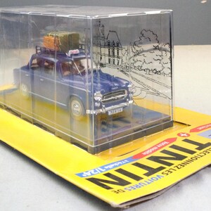 May include: A blue die-cast model car with luggage on the roof, displayed in a clear plastic case. The model is on a yellow cardboard base with the word "TINTIN" printed on it. The car is a vintage style vehicle.