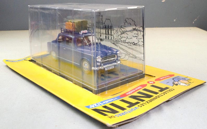 May include: A blue die-cast model car with luggage on the roof, displayed in a clear plastic case. The model is on a yellow cardboard base with the word "TINTIN" printed on it. The car is a vintage style vehicle.