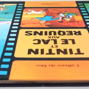 May include: A vintage book titled "Tintin et le Lac aux Requins" with a film strip design. The cover features illustrations of Tintin and Snowy, set against a blue and orange background. The book's spine is yellow.