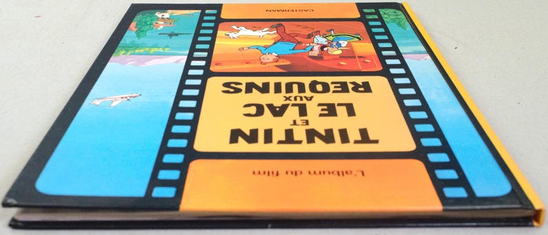 May include: A vintage book titled "Tintin et le Lac aux Requins" with a film strip design. The cover features illustrations of Tintin and Snowy, set against a blue and orange background. The book's spine is yellow.