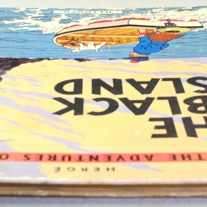 K&ouml;nnte beinhalten: Ein Vintage-Exemplar von "Die schwarze Insel" von Herg&eacute;, aus der Reihe "Tim und Struppi". Das Cover zeigt ein Boot auf dem Wasser, eine felsige Insel und den Titel in gro&szlig;en schwarzen Buchstaben auf gelb-blauem Hintergrund. Der Buchr&uuml;cken ist rot.