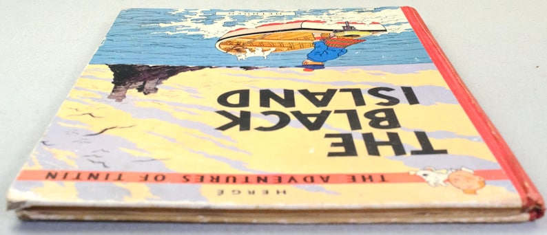 K&ouml;nnte beinhalten: Ein Vintage-Exemplar von "Die schwarze Insel" von Herg&eacute;, aus der Reihe "Tim und Struppi". Das Cover zeigt ein Boot auf dem Wasser, eine felsige Insel und den Titel in gro&szlig;en schwarzen Buchstaben auf gelb-blauem Hintergrund. Der Buchr&uuml;cken ist rot.