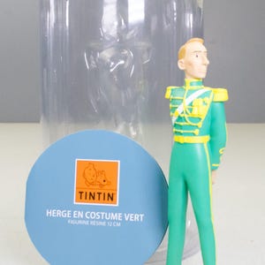 May include: A figurine of a character in a green uniform with yellow accents, standing on a tan base. The figure is approximately 4.7 inches tall. A blue circular label with the Tintin logo and text is in the foreground, with a clear plastic container behind.