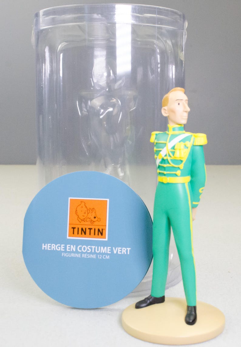 May include: A figurine of a character in a green uniform with yellow accents, standing on a tan base. The figure is approximately 4.7 inches tall. A blue circular label with the Tintin logo and text is in the foreground, with a clear plastic container behind.