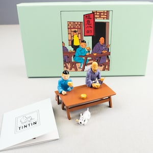 Könnte beinhalten: Ein Set mit drei Tintin-Figuren, die Charaktere aus der Comic-Serie darstellen. Die Figuren sitzen an einem Tisch mit einem weißen Hund. Der Tisch ist braun und hat eine hellbraune Tischplatte. Die Figuren tragen blaue und violette Kleidung. Der Hintergrund ist eine grüne Box mit einem Comic-Bild der Figuren.