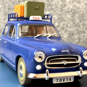 May include: A blue vintage taxi model with a luggage rack holding orange and green suitcases. The car has a "TAXI" sign on top and a license plate that reads "70V14". The model is a Peugeot 403.