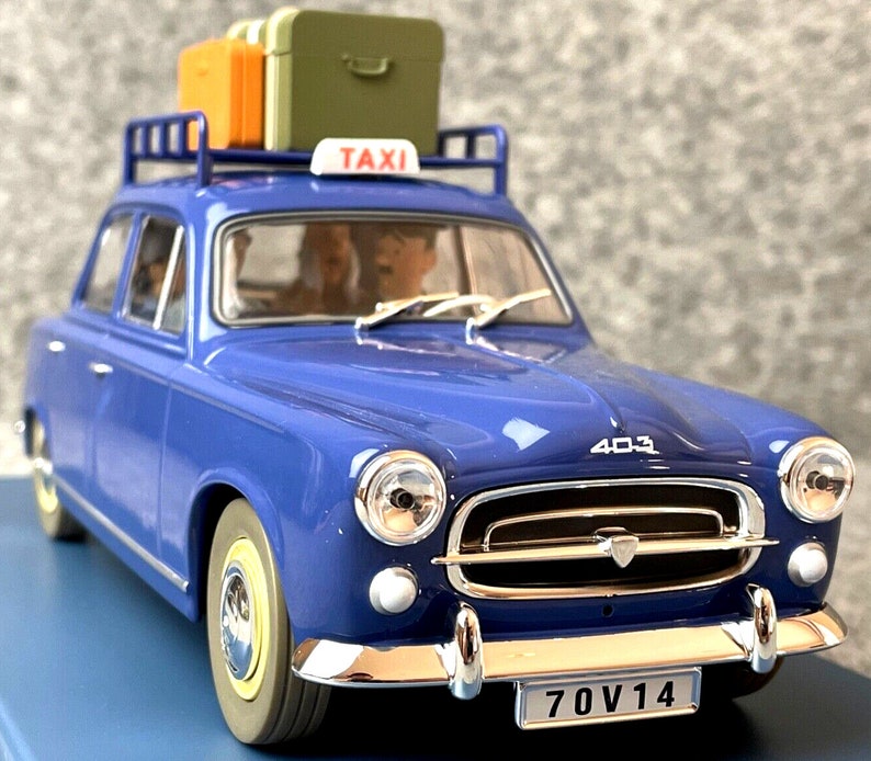 May include: A blue vintage taxi model with a luggage rack holding orange and green suitcases. The car has a "TAXI" sign on top and a license plate that reads "70V14". The model is a Peugeot 403.