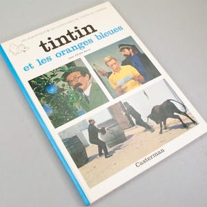 Puede incluir: Un libro vintage titulado "Tintin et les oranges bleues" con una cubierta azul y blanca. La portada presenta ilustraciones de la pel&iacute;cula, incluyendo personajes y escenas. El nombre del editor, Casterman, est&aacute; impreso en la parte inferior derecha.