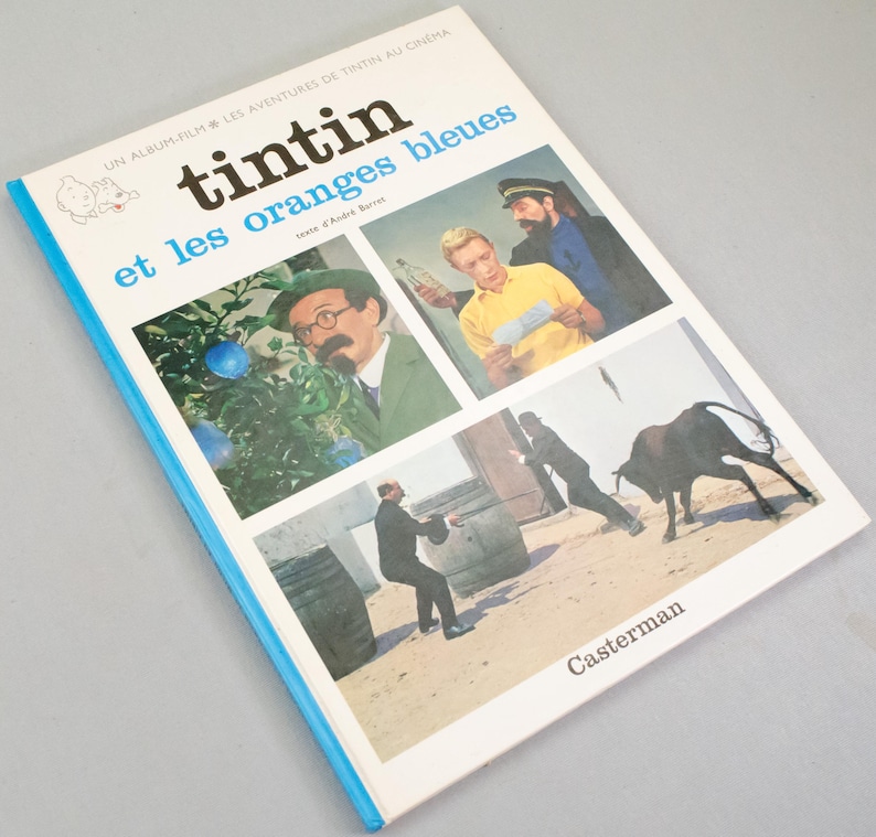 Puede incluir: Un libro vintage titulado "Tintin et les oranges bleues" con una cubierta azul y blanca. La portada presenta ilustraciones de la pel&iacute;cula, incluyendo personajes y escenas. El nombre del editor, Casterman, est&aacute; impreso en la parte inferior derecha.