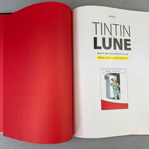 Puede incluir: Un libro abierto con una p&aacute;gina izquierda roja y una p&aacute;gina derecha con el t&iacute;tulo "TINTIN et la LUNE" en negro. La p&aacute;gina incluye una ilustraci&oacute;n de c&oacute;mic de un personaje saludando. El libro est&aacute; sobre una superficie gris.
