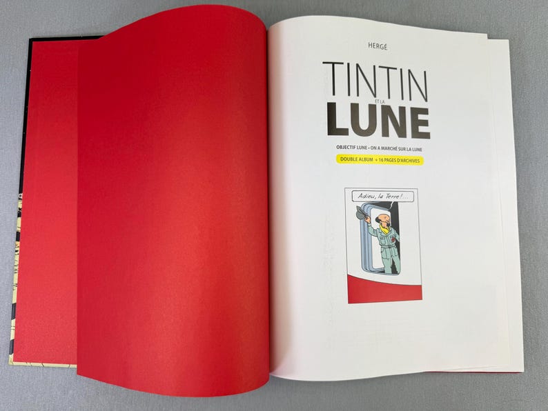 Puede incluir: Un libro abierto con una p&aacute;gina izquierda roja y una p&aacute;gina derecha con el t&iacute;tulo "TINTIN et la LUNE" en negro. La p&aacute;gina incluye una ilustraci&oacute;n de c&oacute;mic de un personaje saludando. El libro est&aacute; sobre una superficie gris.