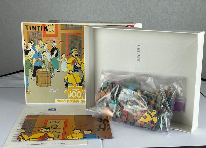 Peut inclure: Un puzzle de 100 pi&egrave;ces repr&eacute;sentant des personnages de la s&eacute;rie de bandes dessin&eacute;es Tintin. La bo&icirc;te du puzzle est ouverte, r&eacute;v&eacute;lant les pi&egrave;ces du puzzle dans un sac en plastique. La bo&icirc;te a une image de Tintin et de ses amis dans un march&eacute; chinois. La bo&icirc;te comprend &eacute;galement le texte "Puzzle 100" et "avec poster gratuit".