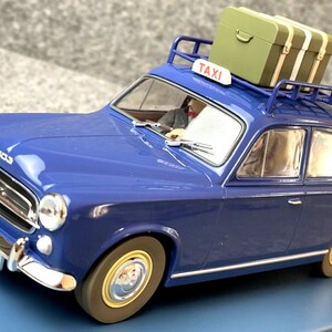 May include: A blue vintage taxi model with a luggage rack and a green suitcase on top. The taxi has a "TAXI" sign and a license plate. The model car has chrome accents and a driver figure inside.