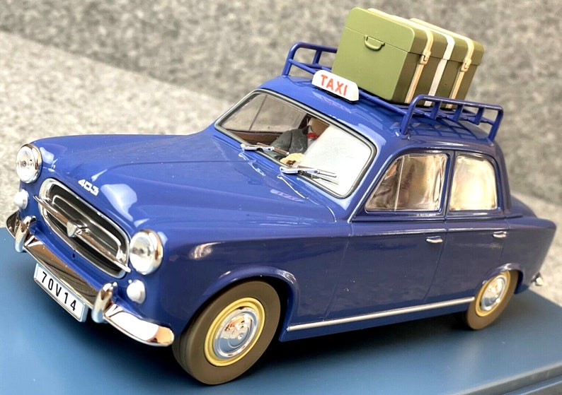 May include: A blue vintage taxi model with a luggage rack and a green suitcase on top. The taxi has a "TAXI" sign and a license plate. The model car has chrome accents and a driver figure inside.