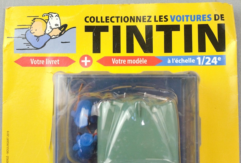 Pu&ograve; includere: Un oggetto da collezione di Tintin confezionato con un modello di veicolo verde in scala 1/24. La confezione &egrave; gialla con testo francese, tra cui "COLLECTIONNEZ LES VOITURES DE TINTIN". Include un'illustrazione di Tintin e Mil&ugrave;.