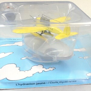 Op de afbeelding: Een geel watervliegtuigmodel in een doorzichtige plastic verpakking, op een blauwe kaart met witte wolkenillustraties. De tekst "L'hydravion jaune - L'&Eacute;toile myst&eacute;rieuse" is zichtbaar. Het model heeft een propeller en drijvers.