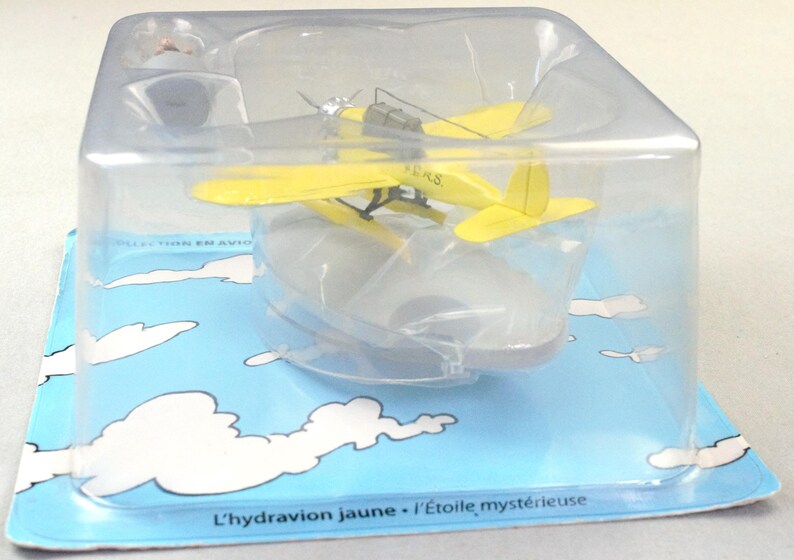 Op de afbeelding: Een geel watervliegtuigmodel in een doorzichtige plastic verpakking, op een blauwe kaart met witte wolkenillustraties. De tekst "L'hydravion jaune - L'&Eacute;toile myst&eacute;rieuse" is zichtbaar. Het model heeft een propeller en drijvers.