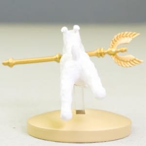 Puede incluir: Figura de perro blanco con un tridente dorado, montada sobre una base circular beige. El tridente tiene una lanza central y dos puntas curvas en forma de hoja. El perro est&aacute; de espaldas al espectador.