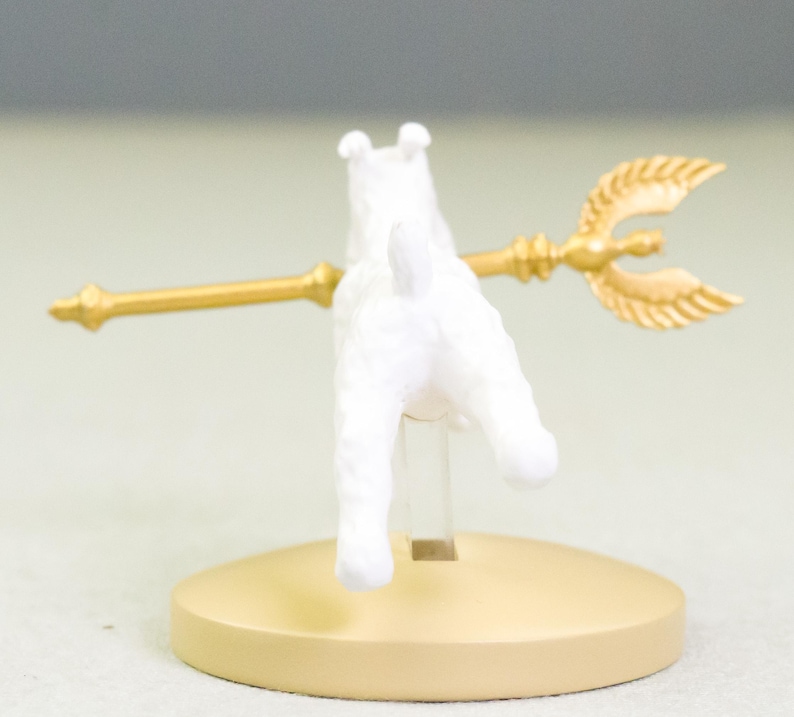 Puede incluir: Figura de perro blanco con un tridente dorado, montada sobre una base circular beige. El tridente tiene una lanza central y dos puntas curvas en forma de hoja. El perro est&aacute; de espaldas al espectador.