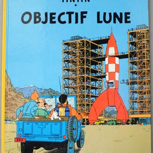 K&ouml;nnte beinhalten: Buchcover von "OBJECTIF LUNE" von Herg&eacute;. Der Hintergrund ist ein blauer Himmel mit einer rot-wei&szlig;en Rakete, einem blauen Jeep mit Figuren und Bauger&uuml;sten. Der Titel ist in gro&szlig;en, fetten Buchstaben.
