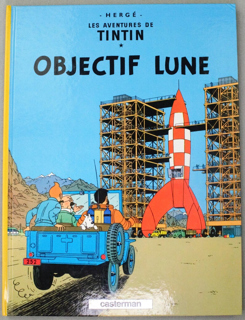 K&ouml;nnte beinhalten: Buchcover von "OBJECTIF LUNE" von Herg&eacute;. Der Hintergrund ist ein blauer Himmel mit einer rot-wei&szlig;en Rakete, einem blauen Jeep mit Figuren und Bauger&uuml;sten. Der Titel ist in gro&szlig;en, fetten Buchstaben.